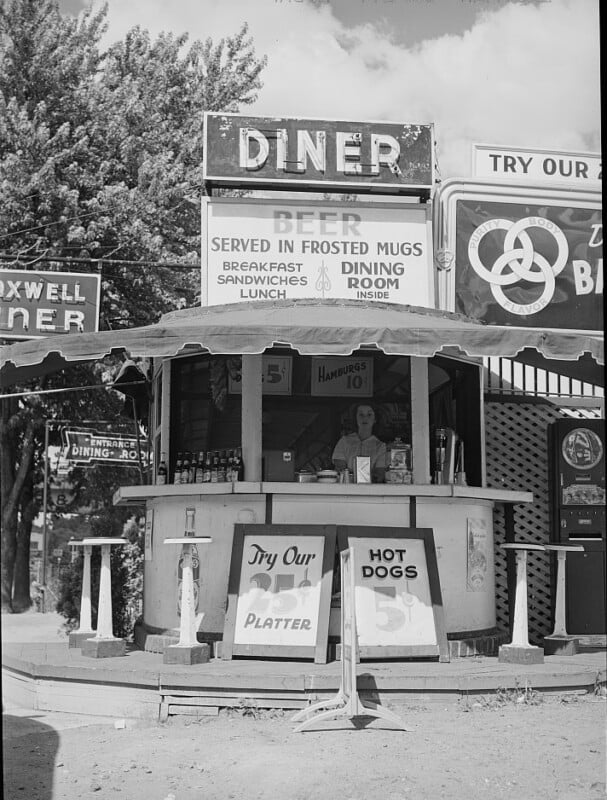 A vintage outdoor diner stand with signs advertising beer, hamburgers for 10 cents, and hot dogs. A server stands inside, and trees and additional signage are visible in the background.