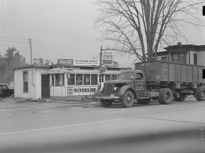 Black-and-white photo of a small roadside diner called “Riverside Diner” with Coca-Cola signs. A large coal truck is parked in front. Leafless tree stands beside the building; empty road and some buildings are in the background.