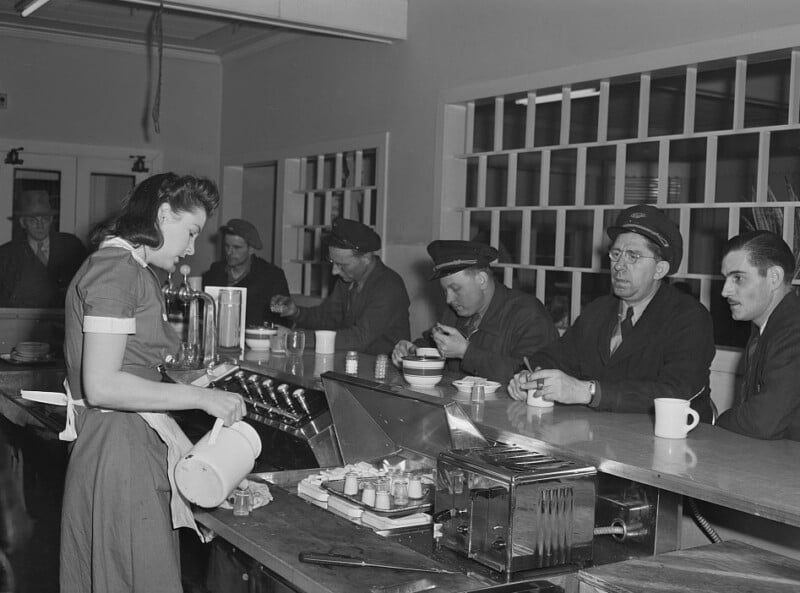 A waitress pours coffee behind a diner counter as several men, some wearing uniforms and hats, sit on stools eating and drinking. The scene appears to be from the mid-20th century.