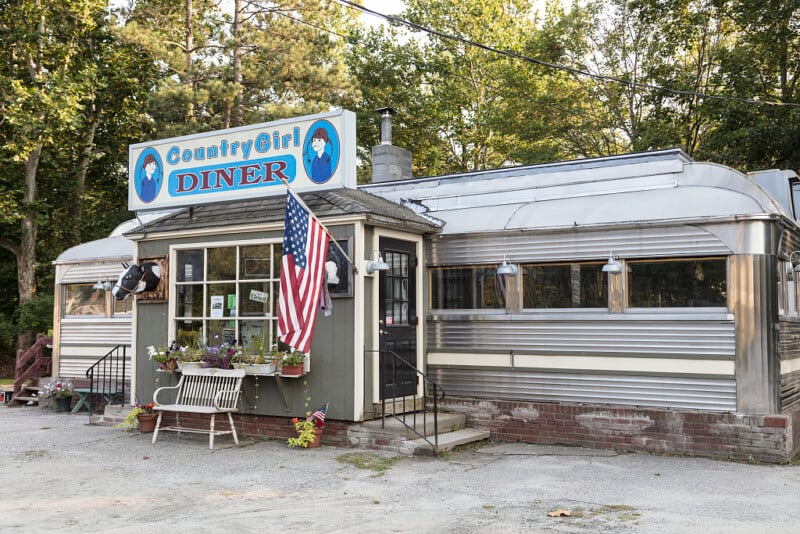 A small, retro-style diner named "Country Girl Diner" with a blue sign, an American flag outside, silver metal siding, and flowering plants near the entrance, surrounded by trees.