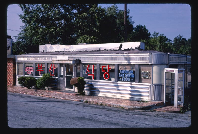 A silver diner with large Korean and English signs advertising “American and Korean Food.” There's a phone booth outside and trees in the background on a sunny day.