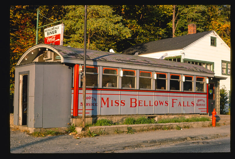 A vintage diner named "Miss Bellows Falls" with red and white signage, large windows, and a Coca-Cola sign sits on a street corner, surrounded by trees and a white house in the background.