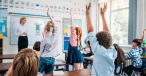 A group of young students in a classroom raise their hands while a teacher stands at the front near a whiteboard. The room is bright with large windows and colorful educational decorations.