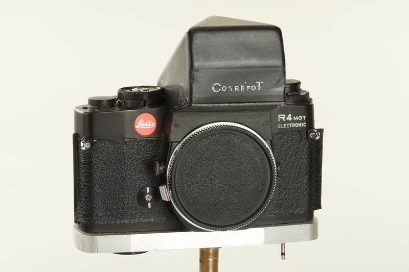 A vintage Cosrefot R4 MOT Electronic film camera with a black body, textured grip, and no lens attached, displayed on a metal stand against a plain background.