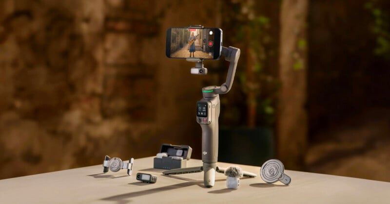 A smartphone mounted on a DJI handheld gimbal is positioned on a table, surrounded by various camera accessories, with a blurred outdoor background.