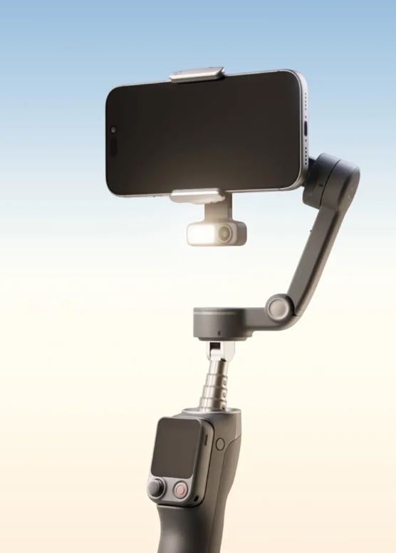 A black smartphone mounted on a handheld stabilizer gimbal with an attached small LED light, set against a gradient blue and beige background.
