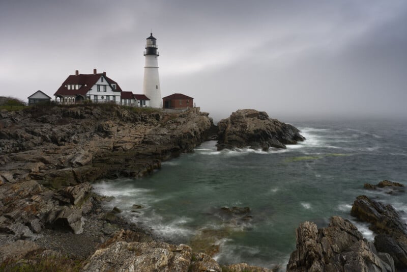 A white lighthouse with an attached red-roofed building stands on rocky cliffs by the ocean under a cloudy, misty sky. Waves crash against the rocks, and the sea appears turbulent.