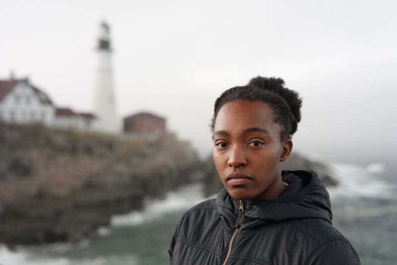 A person wearing a black jacket stands near a rocky shoreline with a lighthouse and buildings visible in the misty background. They are looking calmly at the camera.