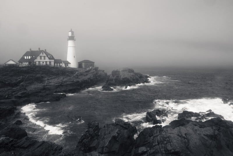 A black-and-white photo of a lighthouse on a rocky coastline with waves crashing against the shore and mist or fog in the background, creating a moody and atmospheric scene.