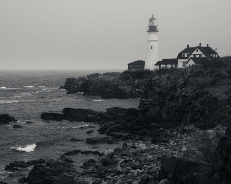 A black-and-white photo of a lighthouse and adjacent buildings on a rocky coastline, overlooking a calm, misty sea under an overcast sky. Waves gently crash against the rocks in the foreground.