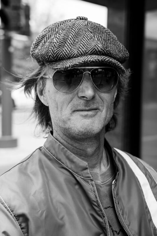 Black and white photo of a person wearing a herringbone flat cap, aviator sunglasses, and a shiny jacket, looking directly at the camera with a neutral expression.