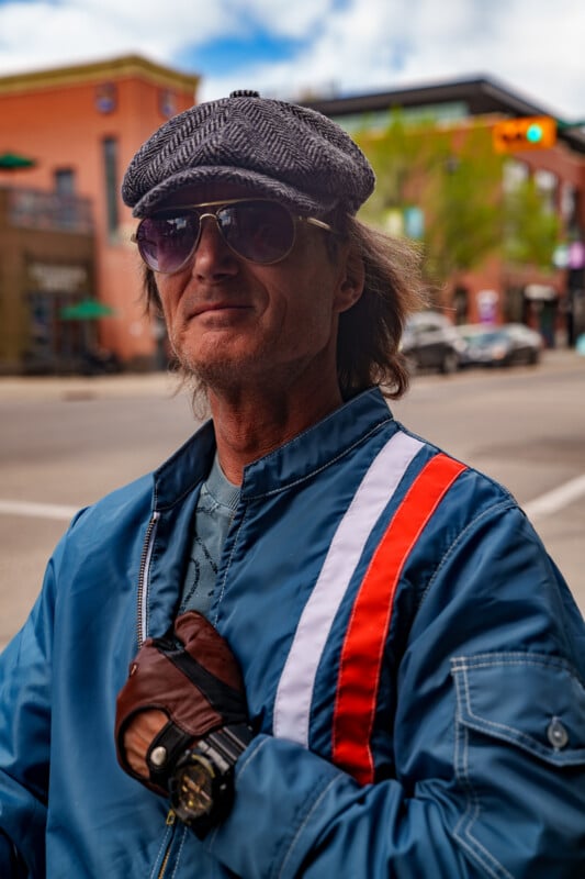 A man wearing a flat cap, sunglasses, fingerless gloves, and a blue jacket with red and white stripes stands on a city street, looking confidently at the camera.