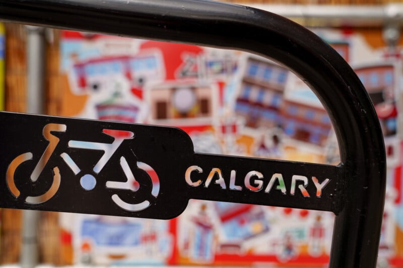 A black metal bike rack with a bicycle symbol and the word "Calgary" cut out, set against a colorful, blurry mural background.