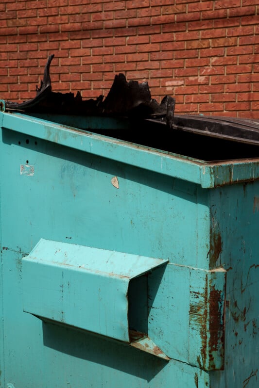 A turquoise dumpster with a partially melted black lid sits in front of a red brick wall. The dumpster is weathered, showing rust and scratches.