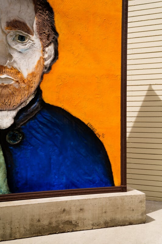 A vibrant mural of a bearded man with reddish hair, wearing a blue coat, is partially shown against a bright yellow background; the scene is cut off on the right and shadow falls on the wall nearby.