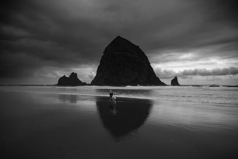 A person walks along the wet sand near the shoreline, with Haystack Rock rising dramatically from the ocean under a cloudy, moody sky at Cannon Beach. The large rock and clouds are reflected in the water.