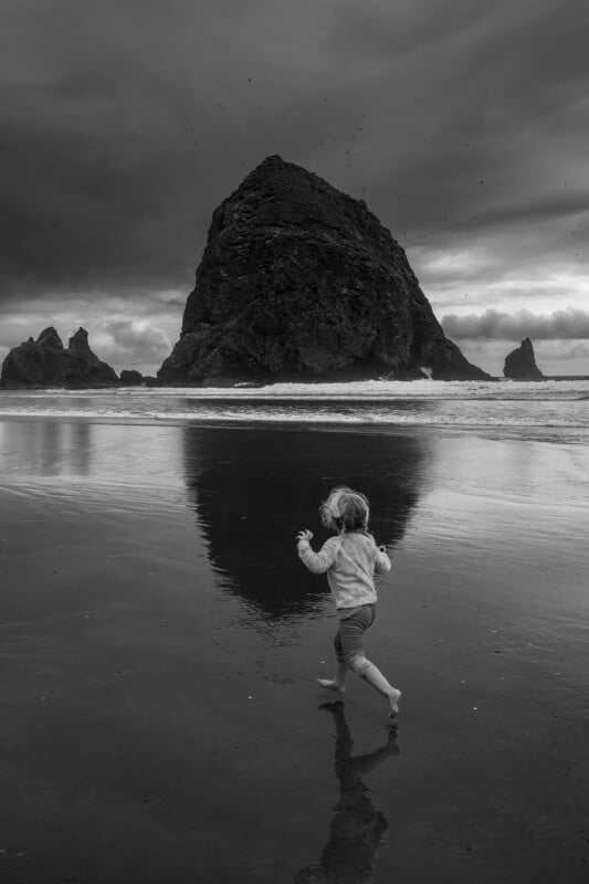 A young child runs barefoot on a wet beach toward a large, dark sea stack under a cloudy sky, with the rock and clouds reflected in the shallow water.
