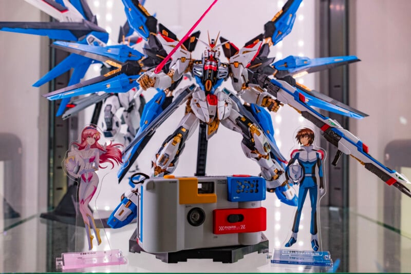 A display case featuring a large, detailed Gundam model with blue and white armor, two anime character standees on either side, and a retro-style camera in front of them. Bright lights illuminate the scene.
