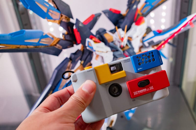 A hand holds a colorful toy-like camera with yellow, blue, and red accents in front of a detailed mecha model with blue, white, and gold armor and wing-like extensions.