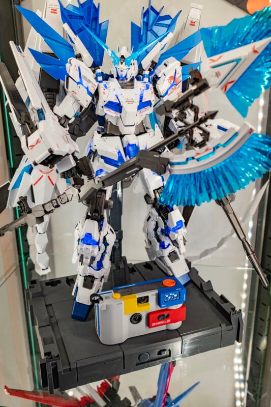 A detailed white and blue Gundam model with large wings and weapon stands on a display base. In front of it is a compact yellow, white, and red reusable film camera on the glass shelf.