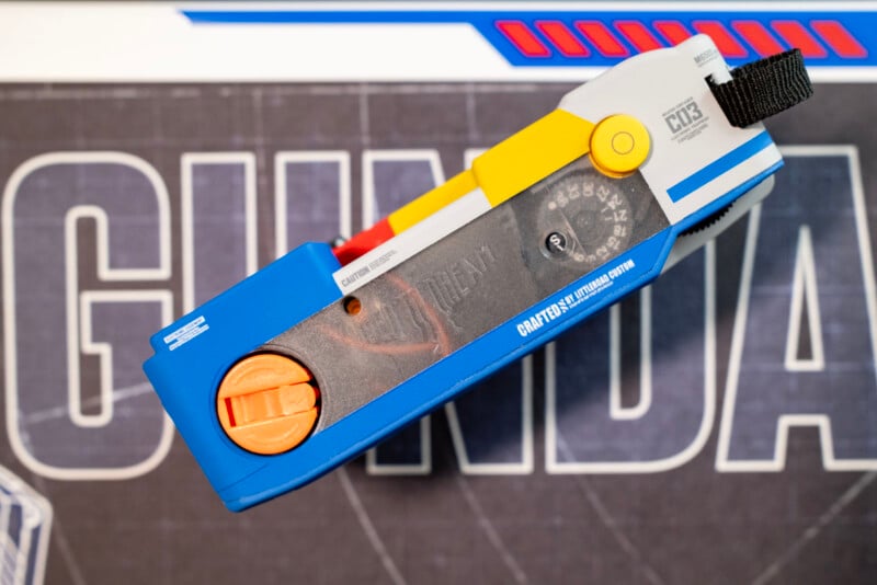 A close-up of a colorful plastic device labeled “CRAFTED Universal Detail” placed on a surface with the word “GUNDAM” in bold, white letters. The device features blue, orange, yellow, and gray components.