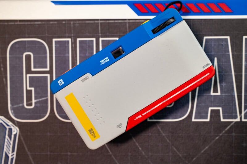 A rectangular computer keyboard with a striking design in blue, yellow, red, and white, inspired by the Gundam anime, placed on a mat with bold graphics and text. The keyboard features a side-mounted scroll wheel.