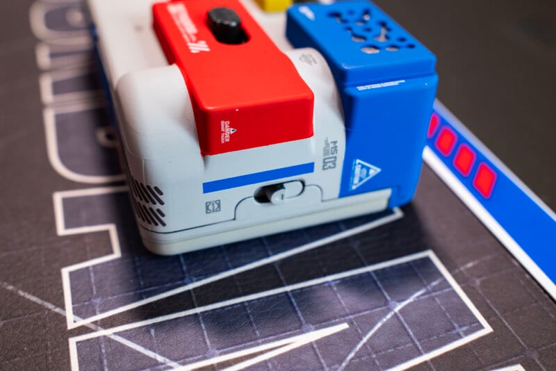 Close-up of a small, colorful robot with red, blue, and white parts on a black mat with white lines and geometric patterns. A blue strip with red rectangles is visible on the right side.