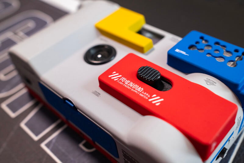 Close-up of a colorful toy-like camera with red, blue, yellow, and gray parts, featuring a prominent black dial and lens on a dark surface with white text in the background.
