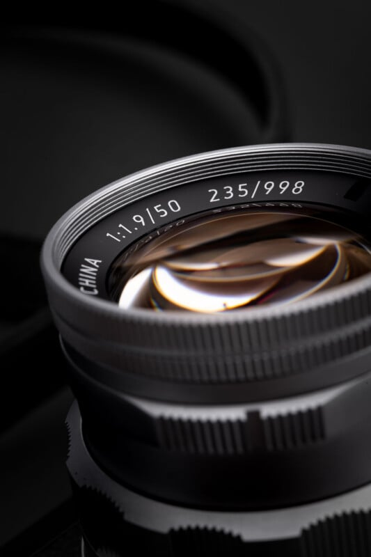 Close-up of a camera lens showing reflections in the glass, with engraved details "1:1.9/50 235/998" visible on the lens rim. The background is dark and out of focus.