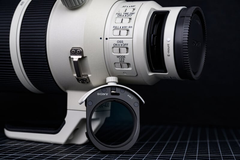 Close-up of a Sony telephoto camera lens with a detachable filter holder placed in front, showing detailed control switches and buttons on the lens body, all set on a grid-patterned black surface.