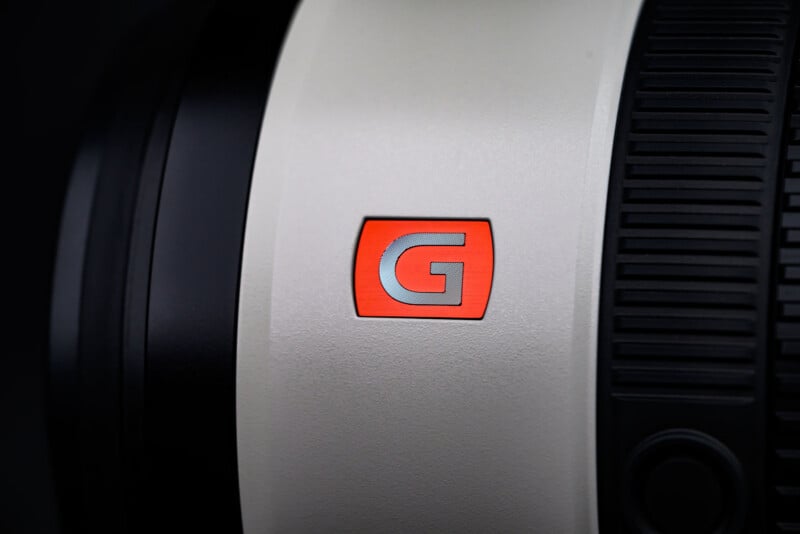 Close-up of a camera lens with a red "G" logo on a silver background, indicating it's a Sony G Master lens. The focus ring is visible on the right side of the image.