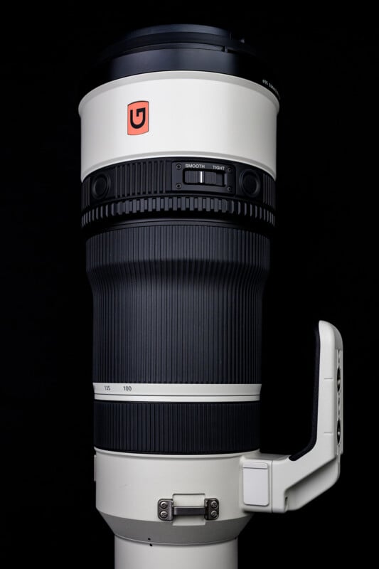A large white telephoto camera lens with a black focus ring, a tripod mount, and a red logo on the upper barrel, set against a black background.
