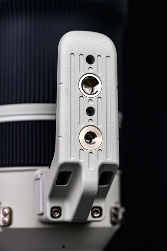 Close-up of a gray, textured tripod collar foot with two metal mounting sockets and screws, attached to a large camera lens against a dark background.