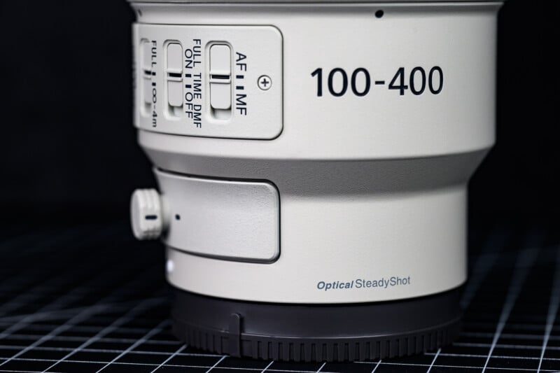 Close-up of a camera lens showing focus mode switches, the numbers "100-400," and the label "Optical SteadyShot" on a grid-patterned surface with a black background.