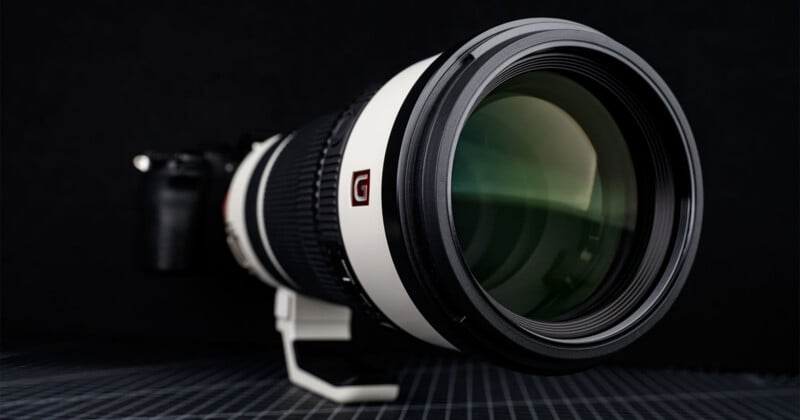 A close-up of a large black and white professional camera with a telephoto lens, featuring a prominent glass element and a sturdy tripod mount, set against a dark background.