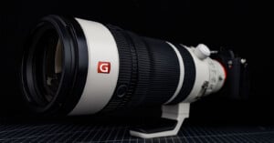 A large professional telephoto camera lens with a white body, black rubber grip, and red "G" logo is mounted on a camera, placed on a grid-patterned surface against a dark background.
