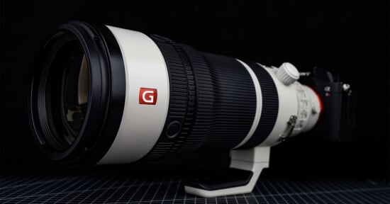 A large professional telephoto camera lens with a white body, black rubber grip, and red "G" logo is mounted on a camera, placed on a grid-patterned surface against a dark background.