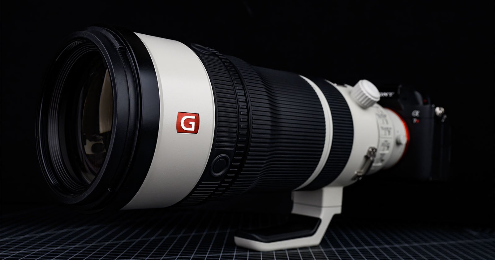 A large professional telephoto camera lens with a white body, black rubber grip, and red "G" logo is mounted on a camera, placed on a grid-patterned surface against a dark background.