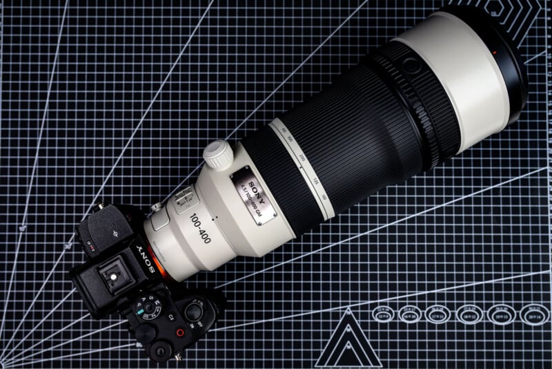 Sony camera attached to a large 100-400mm telephoto zoom lens, placed on a black and white grid cutting mat with measurement lines and geometric shapes.