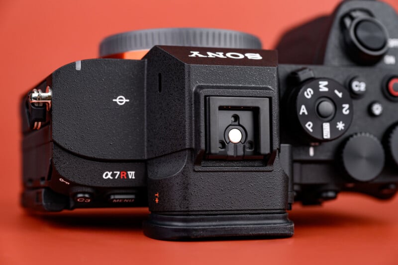 Close-up of the top of a Sony α7R VI mirrorless camera, showing the mode dial, hot shoe, and part of the lens mount against a solid orange background.