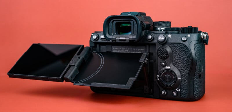 A black digital camera with textured grip is shown from the back against a red background. Its screen is flipped out and angled, revealing buttons and dials on the camera’s rear.