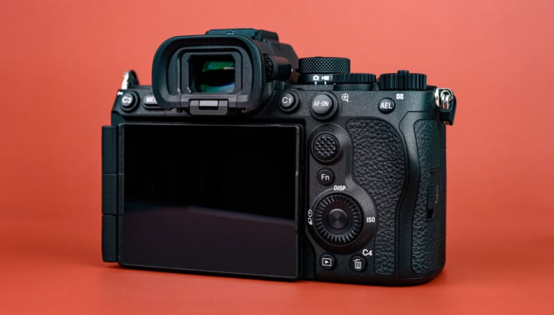 A digital camera with a flip-out screen, various control buttons, and textured grip is displayed against a plain red background. The camera is shown from the back, highlighting the screen and viewfinder.