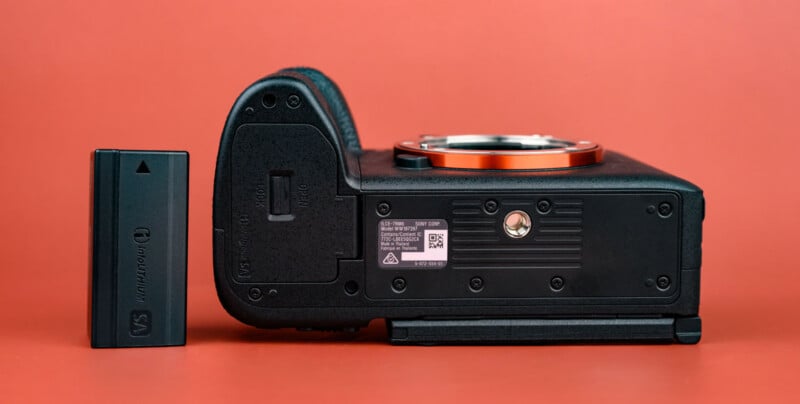 A camera body rests on its side next to a rechargeable battery, both on an orange background. The camera's lens mount and bottom details, including a tripod socket and barcode, are visible.
