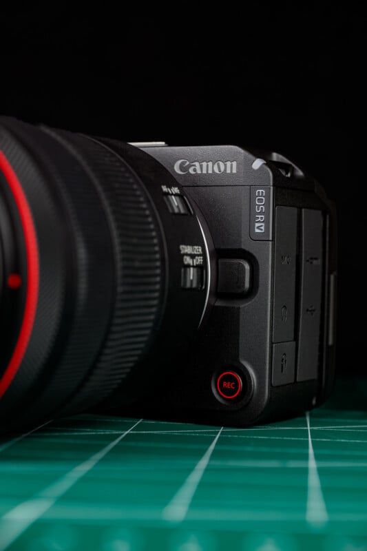A close-up of a Canon EOS R7 camera with a large lens, placed on a green cutting mat with grid lines, photographed against a black background.
