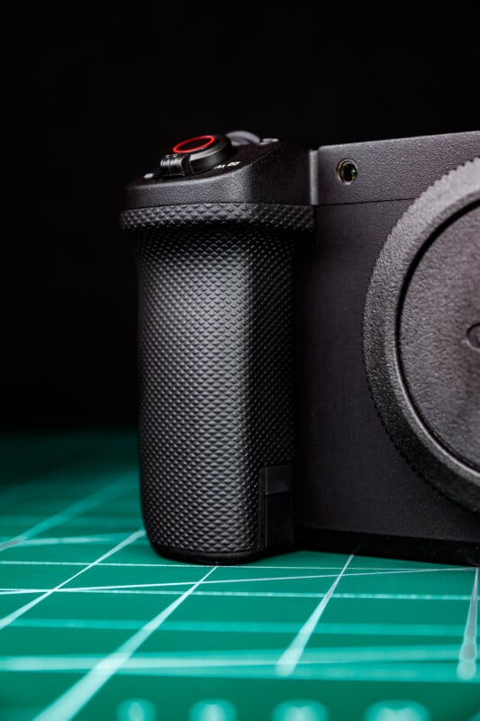 Close-up of a black camera grip with a textured surface, showing part of the camera body, resting on a green cutting mat with white grid lines, against a black background.