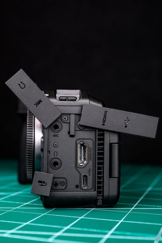 A close-up of a camera's side panel showing open rubber covers revealing ports for microphone, HDMI, USB, and AV out, placed on a green grid surface with a dark background.