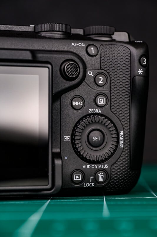 Close-up of the back controls of a digital camera, showing various buttons, dials, and a textured grip next to the LCD screen, placed on a green surface.