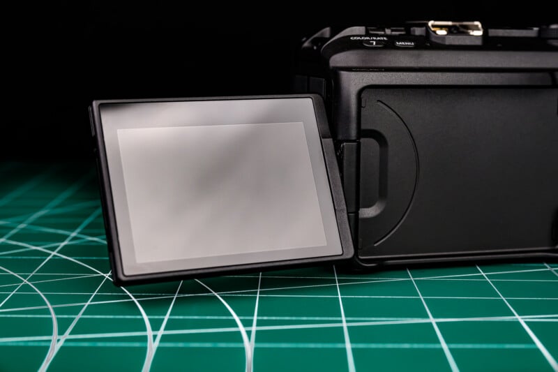 A close-up of a digital camera with its LCD screen flipped out, resting on a green cutting mat with white grid lines, against a dark background.