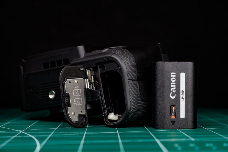 A Canon camera battery grip with its battery compartment open, a Canon LP-E6FP battery beside it, all placed on a green cutting mat with a black background.