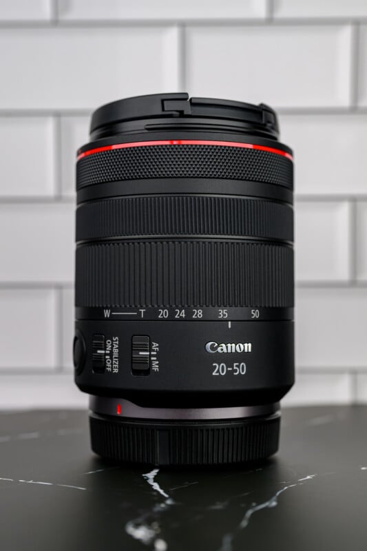 A black Canon 20-50mm camera lens with red accent, shown upright on a dark surface with white tiled background. The lens has visible focus and zoom rings, and switches for stabilizer and autofocus.